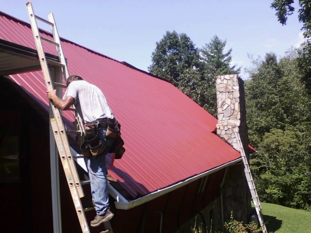 Roofer on a ladder beside a red metal roof and stone chimney — Soffit & Fascia Repair by Roofing Brothers in Fort Valley